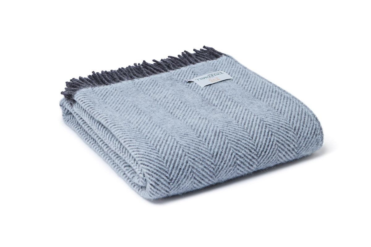 A folded light blue herringbone throw blanket with dark gray fringe and a small white brand tag, shown on a clean white background.
