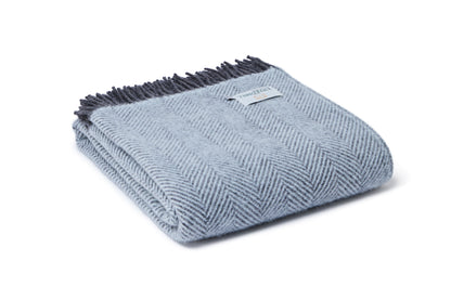 A folded light blue herringbone throw blanket with dark gray fringe and a small white brand tag, shown on a clean white background.