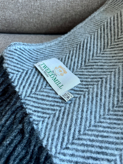 A close-up of a light gray and white herringbone patterned knit blanket with a visible woven label reading Tweedmill.
