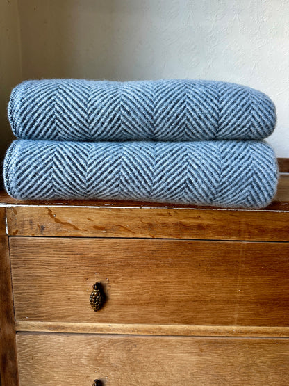 Two folded blue-gray herringbone woven blankets stacked on a rustic wooden chest of drawers against a plain off-white wall.