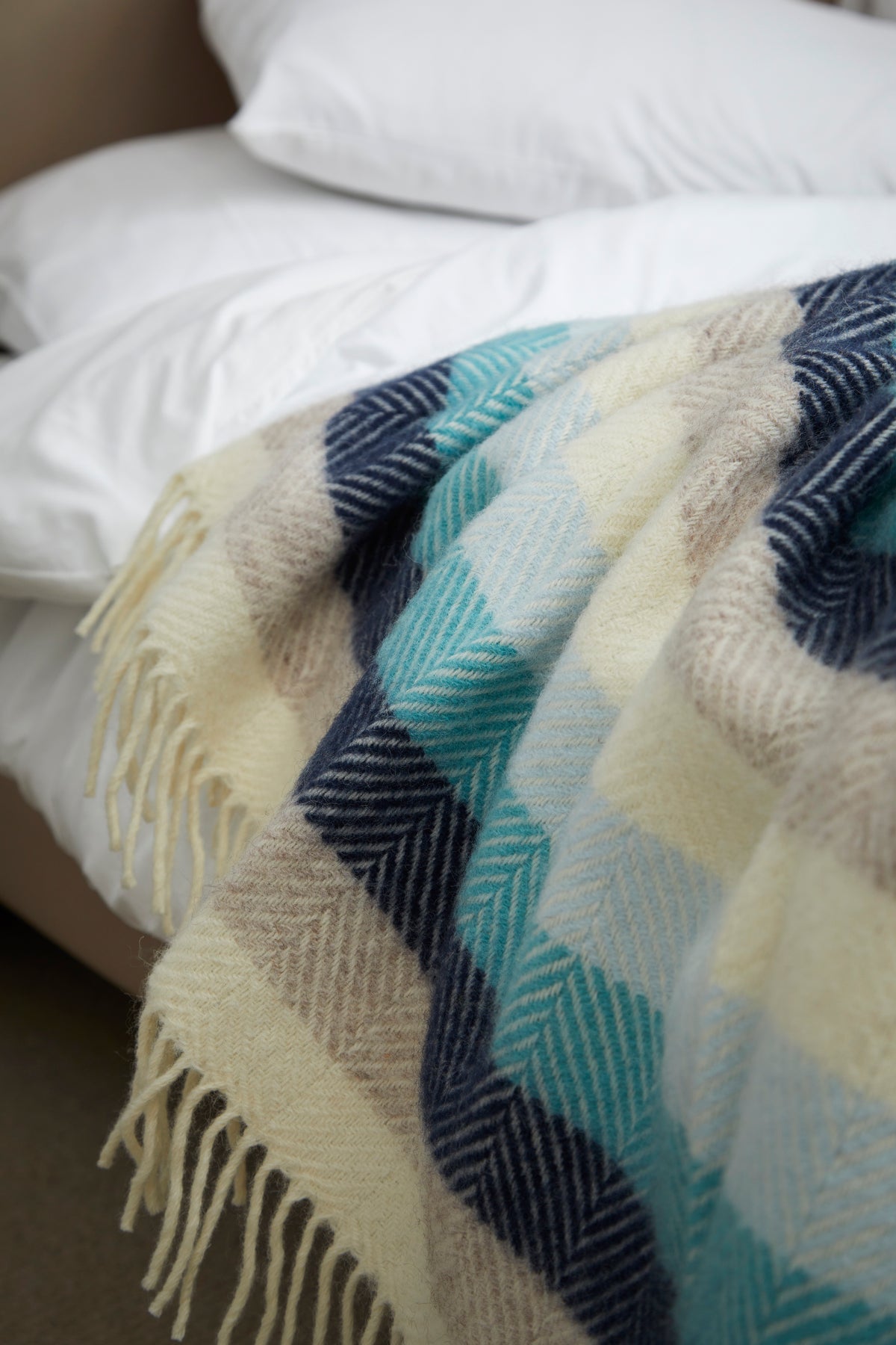 A close-up of a woven throw blanket draped over a bed. The blanket features a large-scale plaid pattern in cream, light blue, navy, and beige, with cream-colored fringe along the bottom edge.