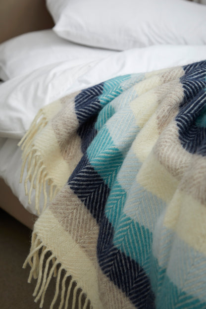 A close-up of a woven throw blanket draped over a bed. The blanket features a large-scale plaid pattern in cream, light blue, navy, and beige, with cream-colored fringe along the bottom edge.