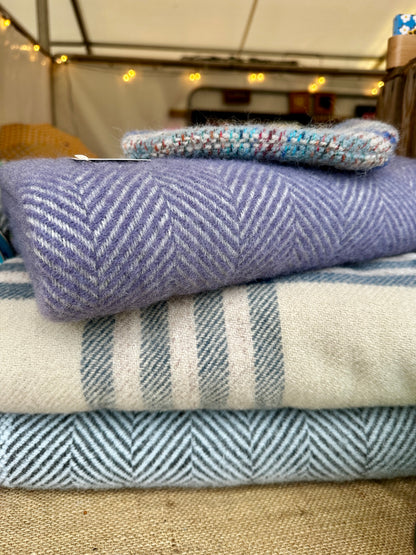 A stack of folded woven textiles, including a lavender-purple herringbone blanket, a cream and blue striped throw, and a light blue herringbone piece, with a small multicolored knit item on top, photographed in a tent-like setting.
